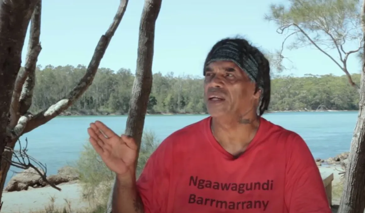 A man wearing a bandana and a red t-shirt looks to the right with his right hand raised. His shirt has the words Ngaawagundi Barrmarrany. In the background are trees, a river and beyond that, a forest of trees.