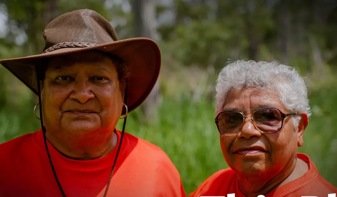 Two Aboriginal women in crimson t-shirts, one wearing a brown brimmed hat and the other with grey hair and wearing sunglasses, look to camera. In the background is green grass and trees.