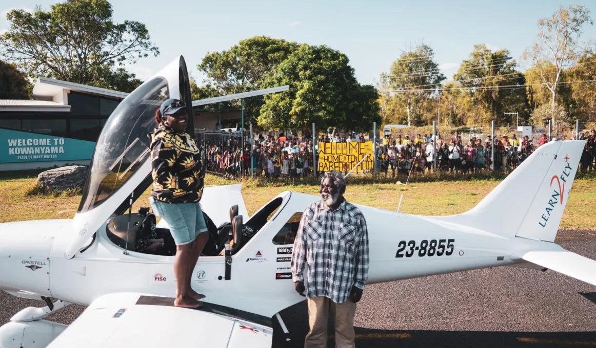 A young Indigenous woman, Tyeisha Clarke, is standing in bare feet on the wing of a light aeroplane, after exiting the cockpit. She is pictured with her father John. The Kowanyama community are standing behind a secure fence away from the airstrip.