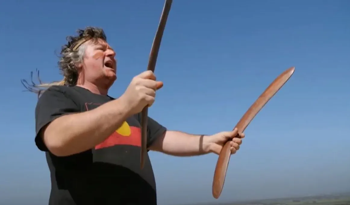 A middle aged man with wavy hair dressed in black shirt with Aboriginal flag on the front holds a boomerang in each hand and is in the attitude of singing as he looks out across the landscape. In the background is blue sky and a strip of green land.