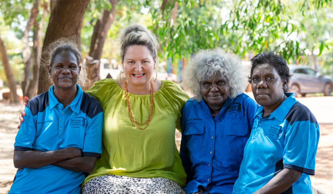 Four women ranging in age from middle-aged to elderly and dressed in different coloured shirts sit in front of trees. In the background are some cars.