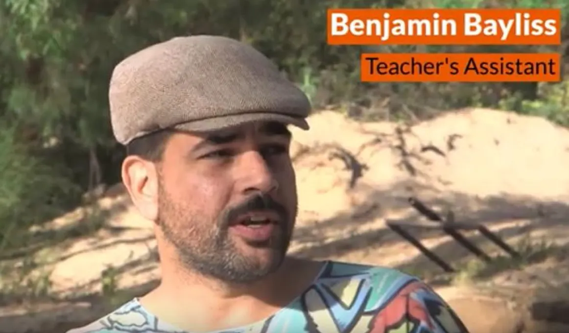 Aboriginal man in cap in front of pile of sand and green shrubbery.