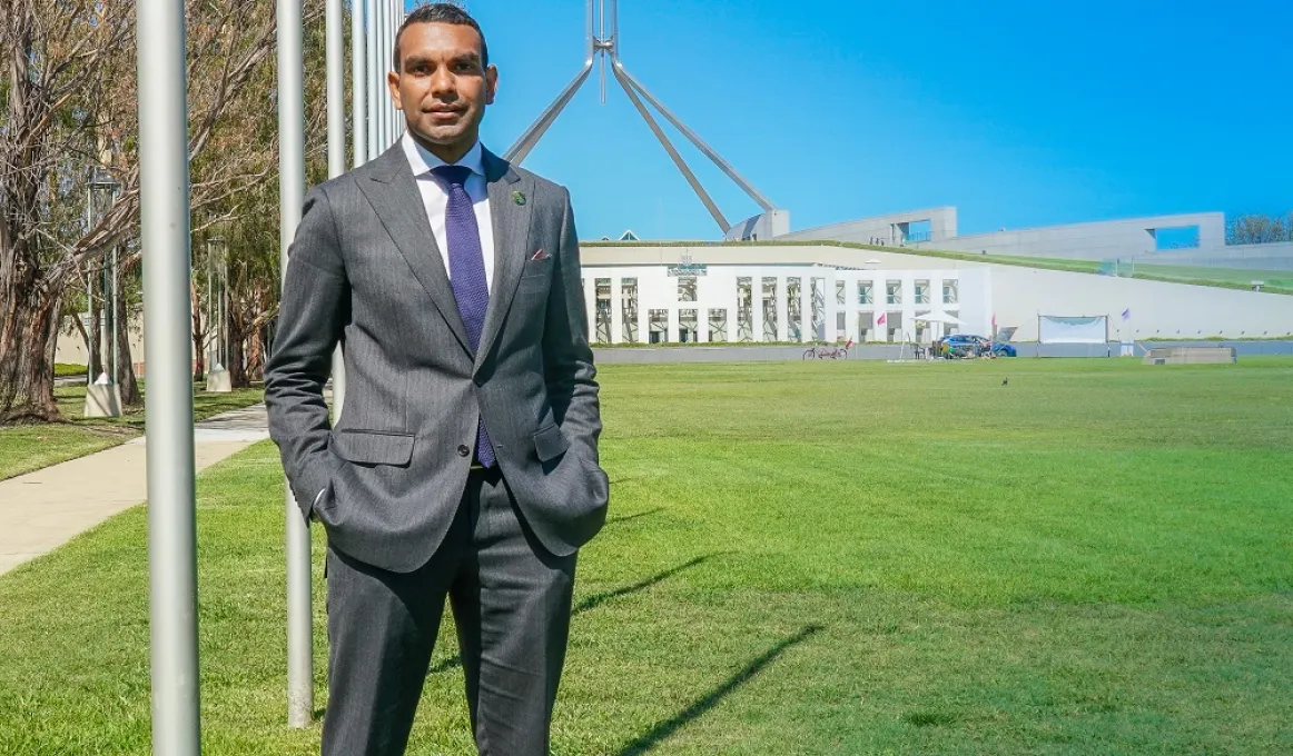 Image of a man wearing a grey suit, standing on the left, facing the camera with Parliament House behind him, a very blue sky and green grass.