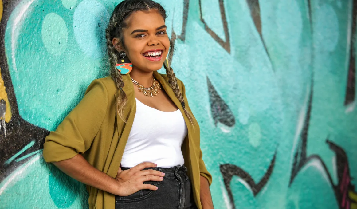 An Indigenous young adult woman, her hair plaited and wearing a green yellow cardigan, white top  and black jeans, leans up against a wall painted black and with an aqua coloured mural.