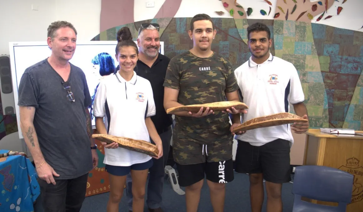 Five people in causal wear stand in a room. Three young adults carry wood objects. Next to them is a blue chair on one side and a table to the other side. Behind them is a white board and colourful wall.