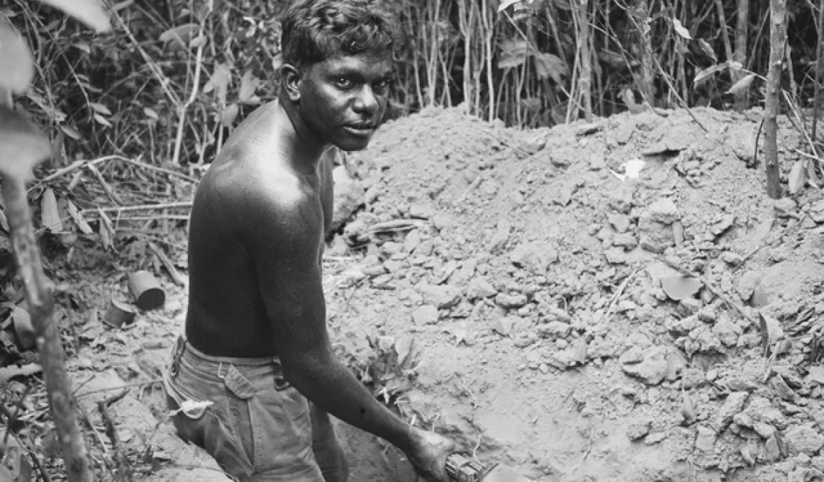 A young man wearing shorts but bare chested holds a shovel in his hand while he stands in a trench. In front of him is a pile of soil and beyond that is thick foliage.