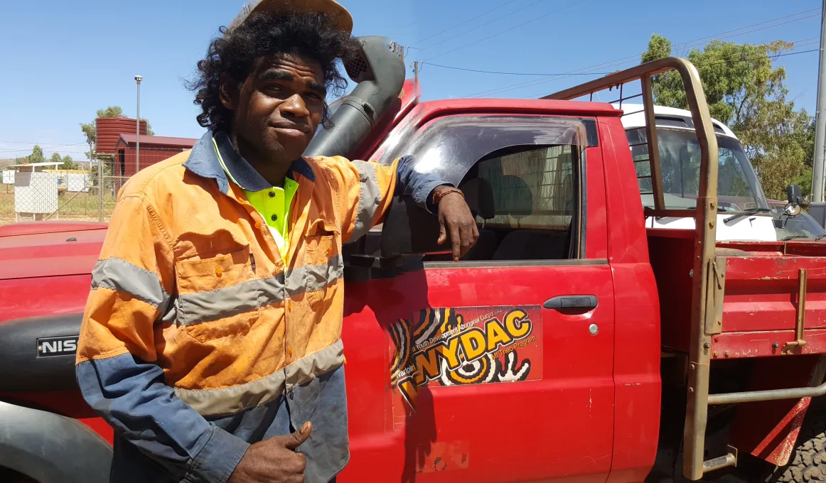 A young Indigenous man dressed in work wear stands in front of a red truck with the letters WYDAC on the cab door. In the background is a white vehicle, trees and buildings.