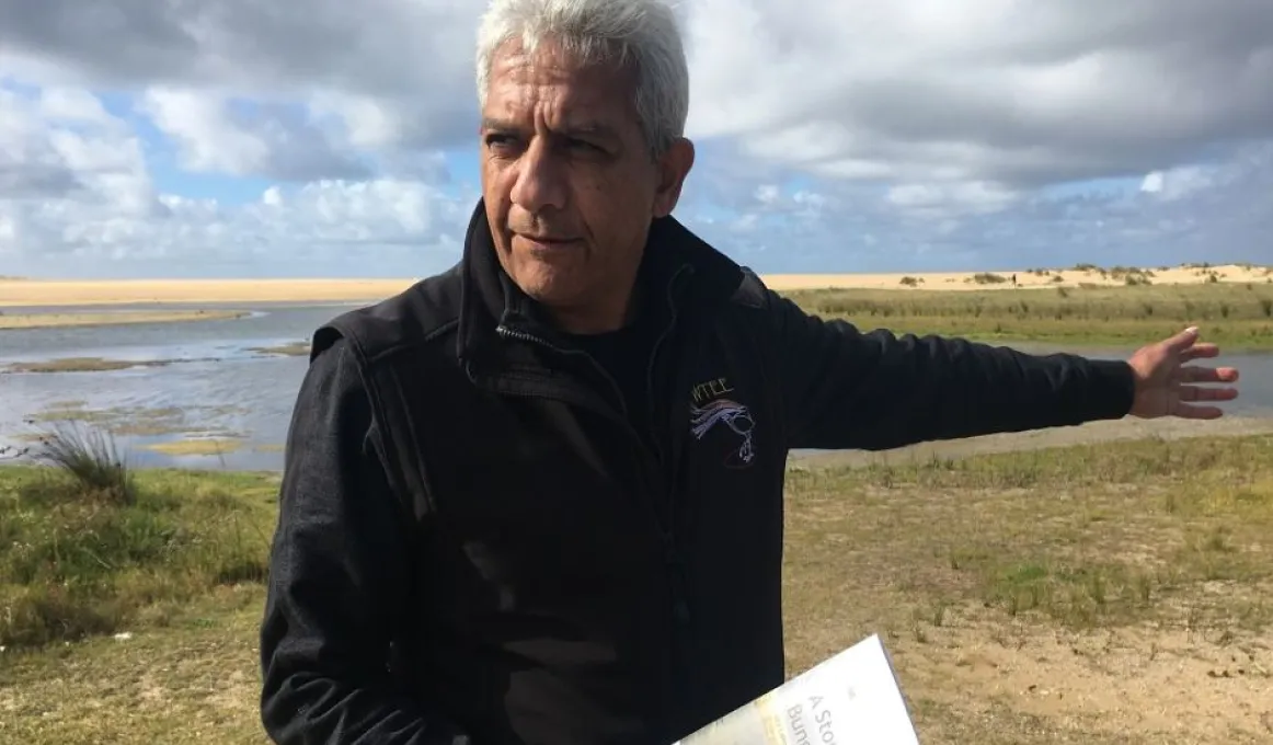 Aboriginal man in dark jacket holds a book in his right hand and points with his left. In the background is grass, water, sand and sky.