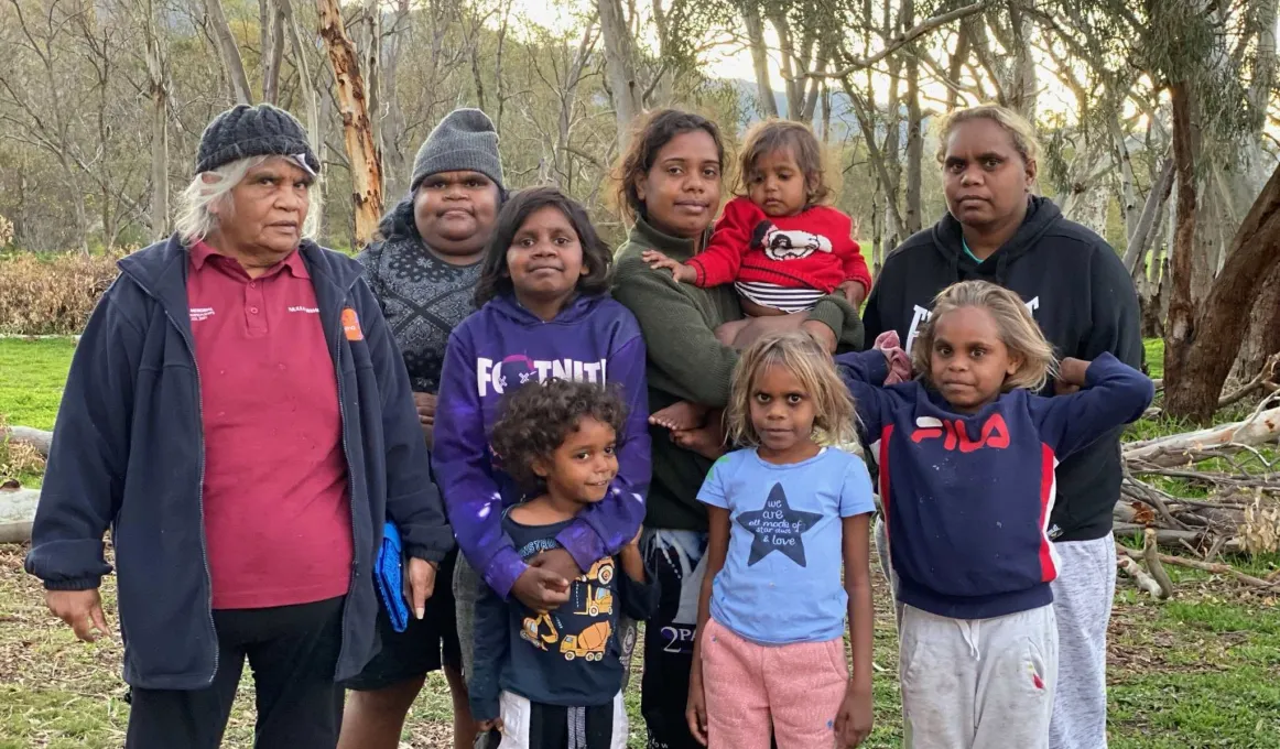 Group of Aboriginal women and children dressed in casual wear stand on a grassy surface. In the background are trees and a hill.