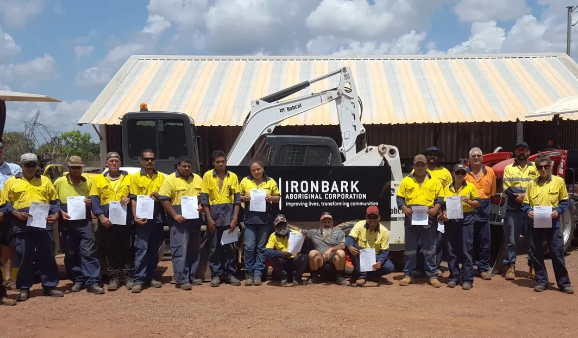 A group of men and women holding certificates while standing in front of heavy machinery and a shed.