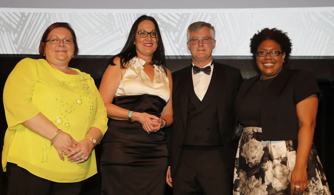 Four adults stand in a row: woman in yellow top, woman in white and black dress, man in tuxedo and woman in black top and black and white mottled skirt. All wear glasses and stand in front of a two toned wall.