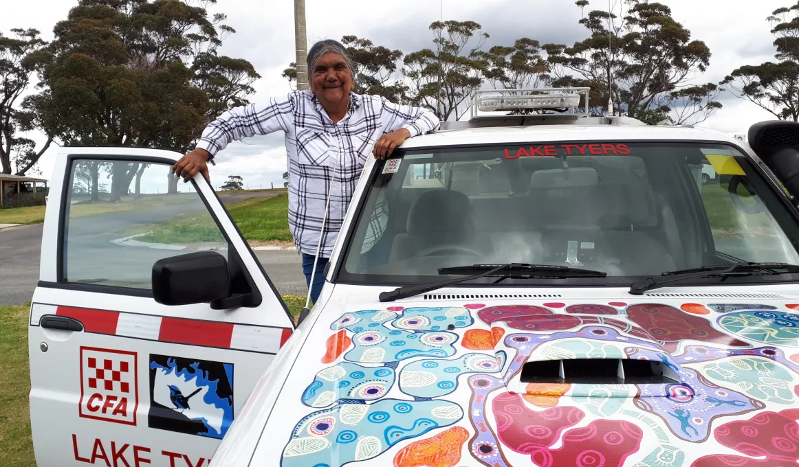 A car with a brightly painted front with the words Lake Tyers CFA written on the vehicle. A woman is standing in the door of the vehicle with the door open and smiling wearing a white and black striped shirt.