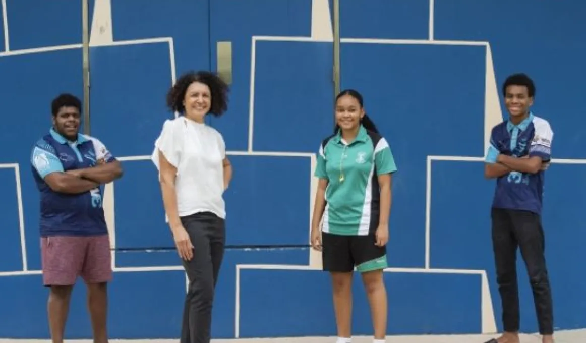 3 youth and an adult woman stand on a carpeted floor. Behind them is a blue wall with white designs.