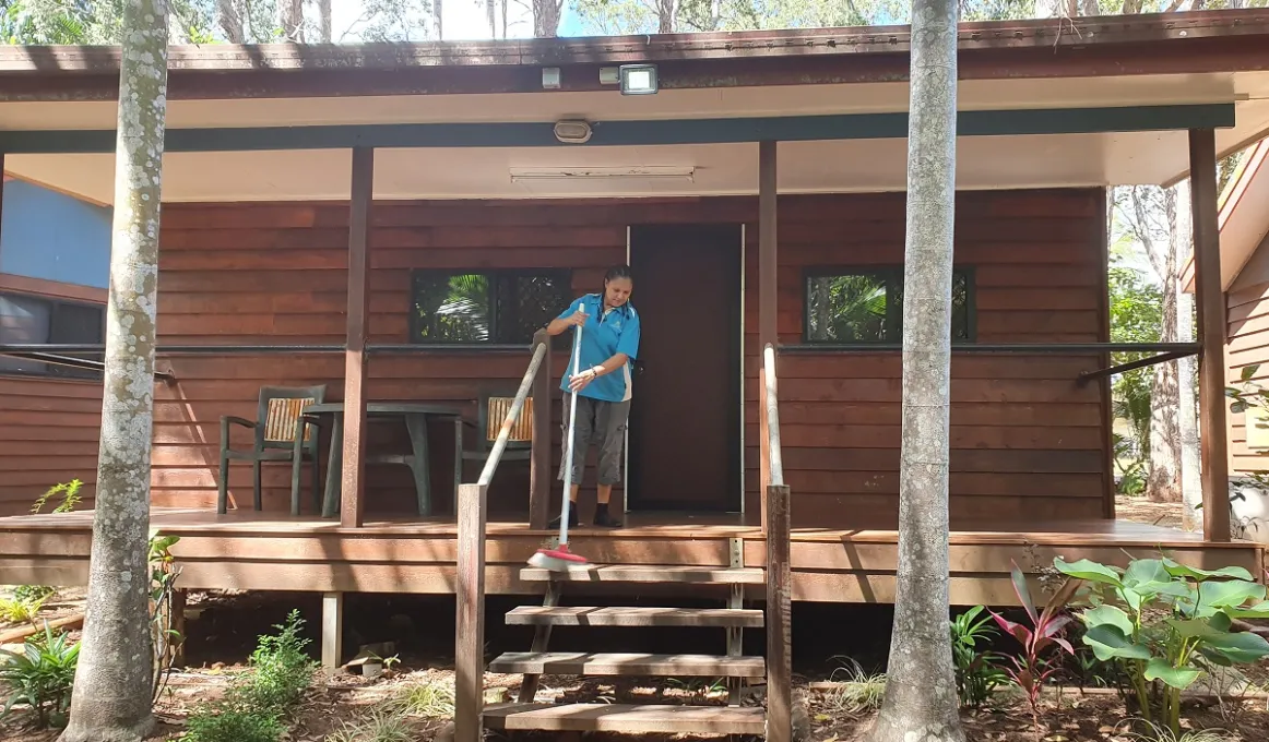 A woman in work wear sweeps the deck in front of a wooden cabin. At the front are two palm trees, some foliage and steps leading up to the cabin entrance. In the background are more trees.