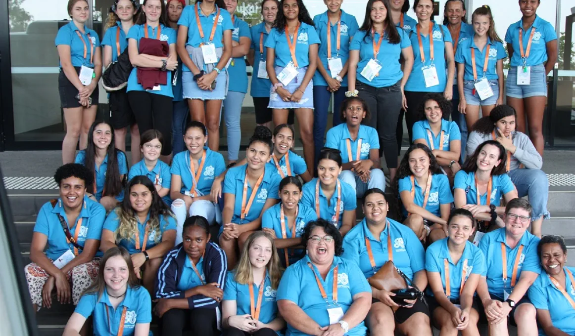 Large group of young Indigenous women sit and stand on several rows of stairs. Most wear a blue shirt.