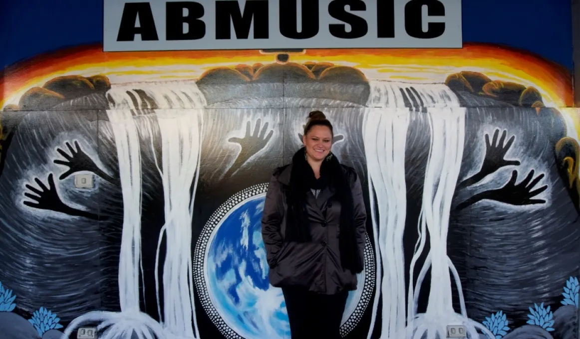 Indigenous woman standing in front of a large grey and white and blue painting of black hands and white waterfalls and the word ABMUSIC at the top.