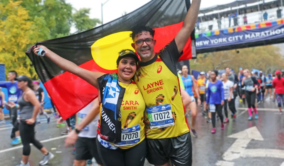 An Aboriginal woman and man stand on a street holding the Aboriginal flag. Behind are many people dressed in running wear crossing a finish line, above which hangs a banner on an overhead walkway.