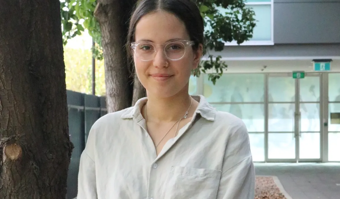 A young woman with dark hair, wearing glasses and a pale shirt stands in front of some trees. In the background and to the right is a building entrance made mainly of glass windows set in metal frames.