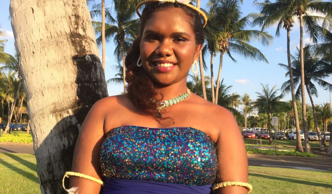 Young Tiwi Island woman in purple dress and headdress standing in front of a palm tree.