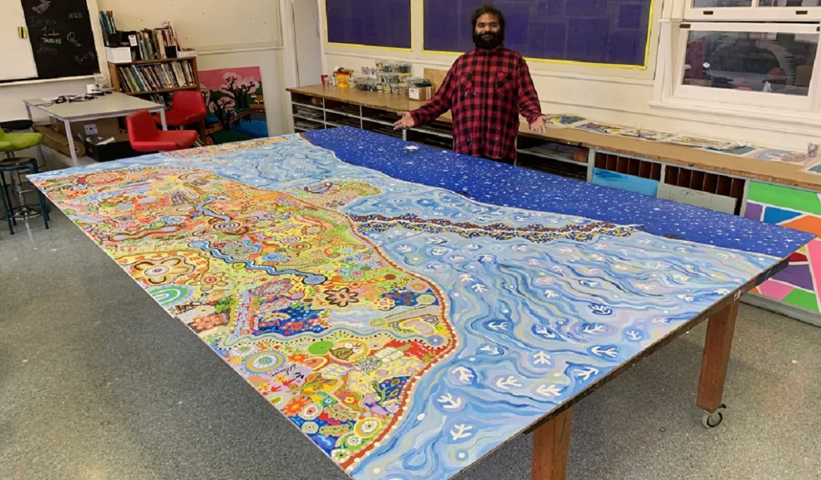 A man wearing a black and red checked shirt is standing in front of a large colourful canvas painting which is on a table.