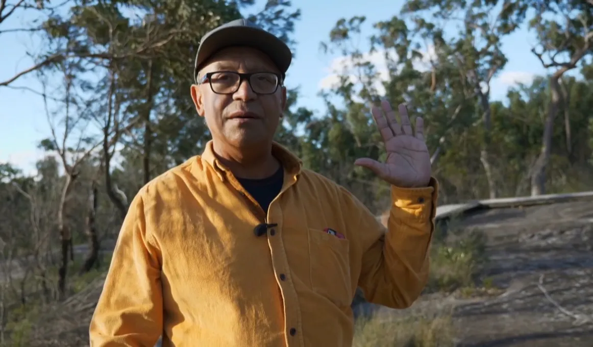 Elderly man in yellow shirt and peaked cap stands with left hand raised. In the background are trees and bare ground.