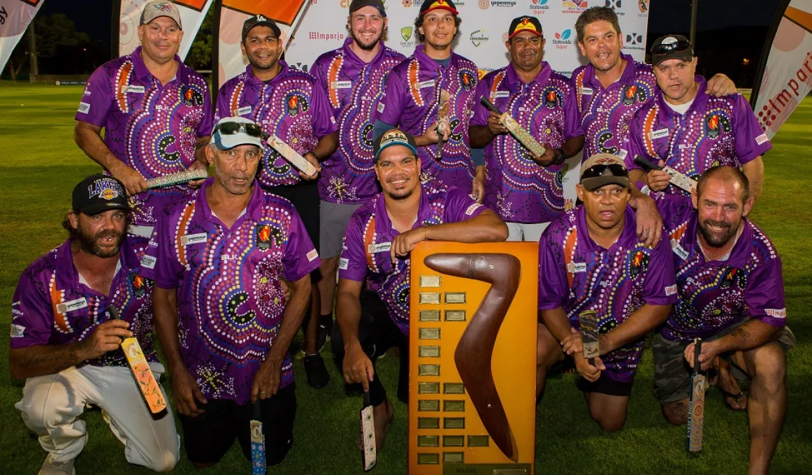 Group of men in purple jerseys featuring Indigenous designs, stand or kneel around a large trophy featuring a boomerang on a large wooden plaque with small metal plates.