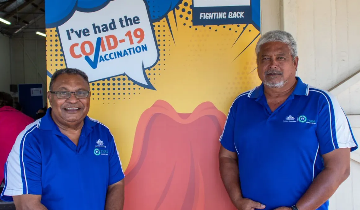 Two men in blue shirts face camera. In the background is a poster featuring the text: hero, I've had the Covid-19 vaccination. Covid-19 Issue #1 fighting back.