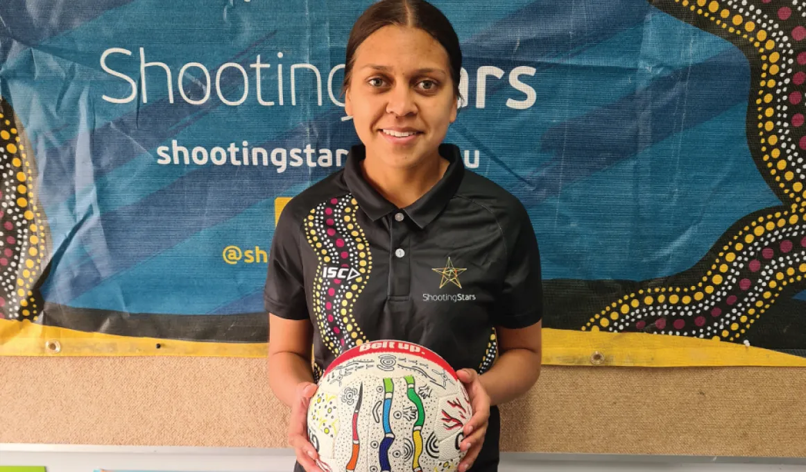 A young woman with her black hair pulled back holds a white ball in her hands. On the ball are traditional Aboriginal designs. In the background is a blue poster with the words Shooting Stars and some more Aboriginal artwork.