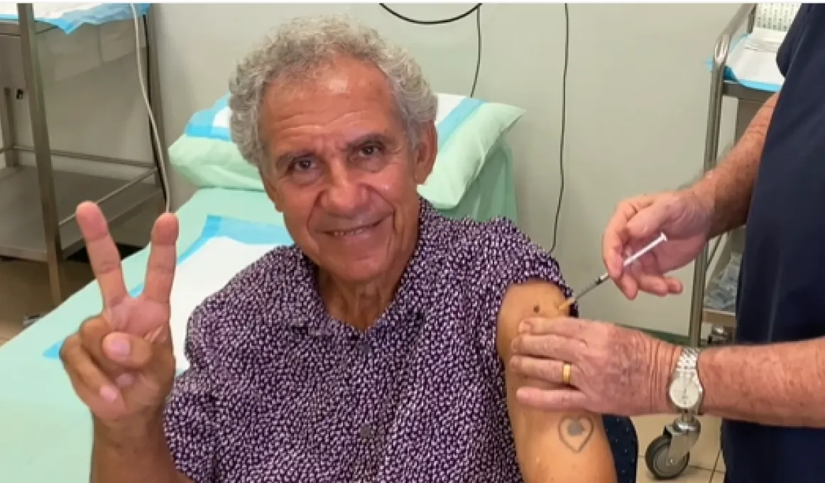 A man in a purple and white spotted shirt is sitting next to a medical bed with two fingers in a peace sign. A second man in blue scrubs is putting a needle in the first man’s arm.