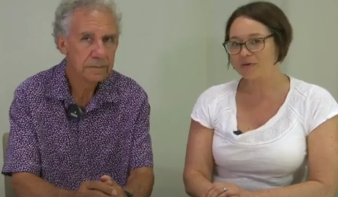 Man and woman set side-by-side at a white desk with a plain grey wall in the background. The man is sat on the left and is wearing a short-sleeve, button-up shirt with a purple pattern. The woman is on the right-hand-side, wearing white t-shirt