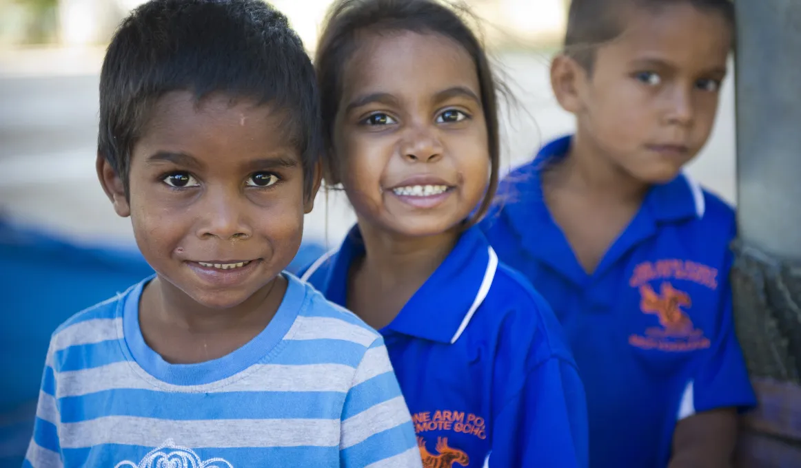 Young Indigenous male child in white and blue striped shirt in front of and to left of similar aged Indigenous girl in dark blue shirt who is in front of another Indigenous boy in same type of shirt.