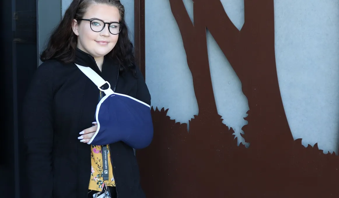 Woman with long dark hair and wearing glasses has her left arm in a blue sling. In the background is a flat brown tree sculpture on a blue wall.