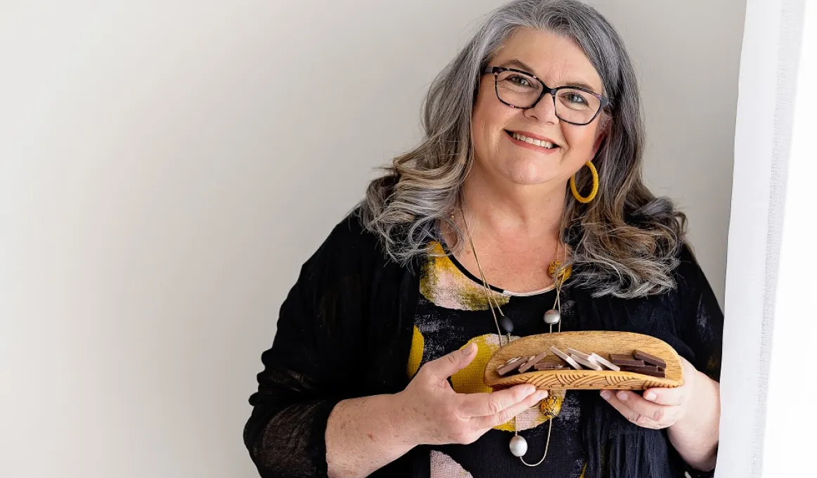 A woman – head tilted to her left, and bearing a broad, friendly smile – cradles an array of chocolates in a coolamon (a traditional Indigenous carrying vessel). She is wearing clothing and jewellery in a mixed palette of bright earth and dark tones.