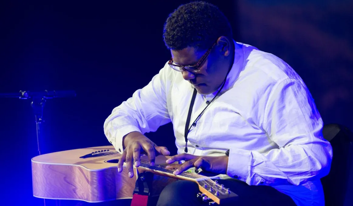 Indigenous man playing a guitar and wearing pale shirt and dark trousers.