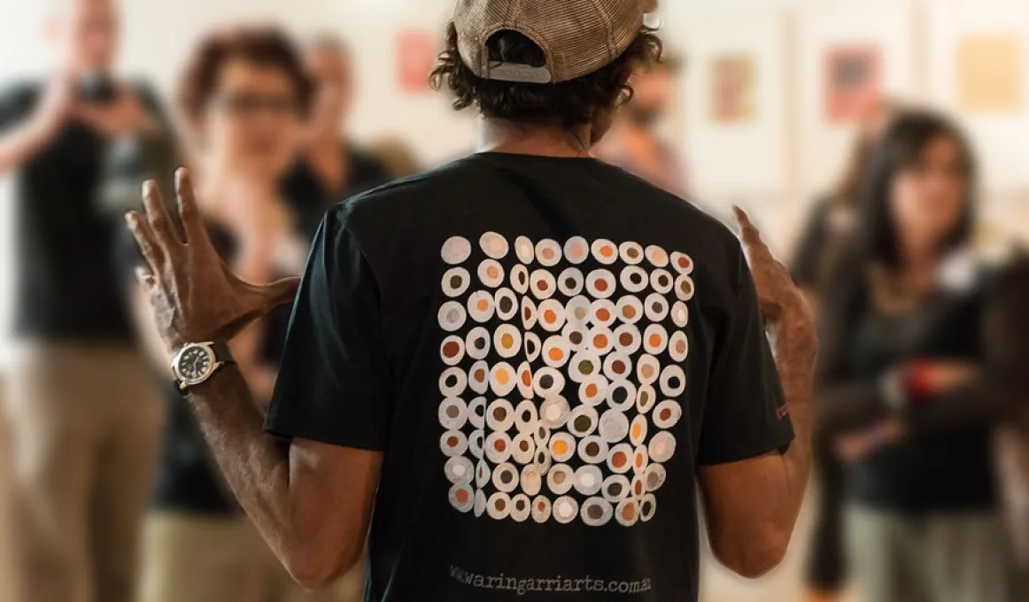 Indigenous man with back to the camera, wearing black t-shirt with Indigenous design on the back. He has his hands raised to shoulder level. In background are group of people looking at him.