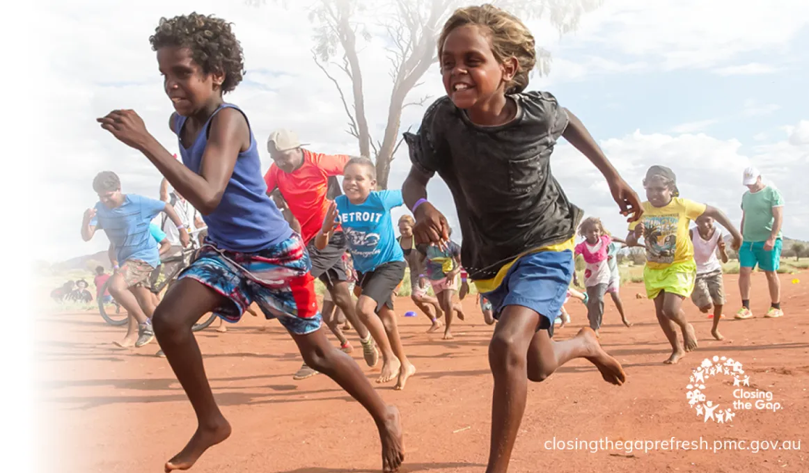 Two Indigenous boys run across red dirt with other children following behind. They are smiling. Closing the Gap logo. closingthegaprefresh.pmc.gov.au