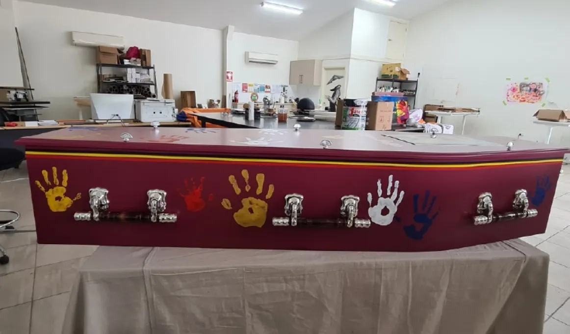 Side view of a redwood coffin with 3 silver coloured handles siting on a bench covered by a brown sheet. There are several hand prints in different colours on the side of the coffin. It is contained in a room and in the background are workshop items.