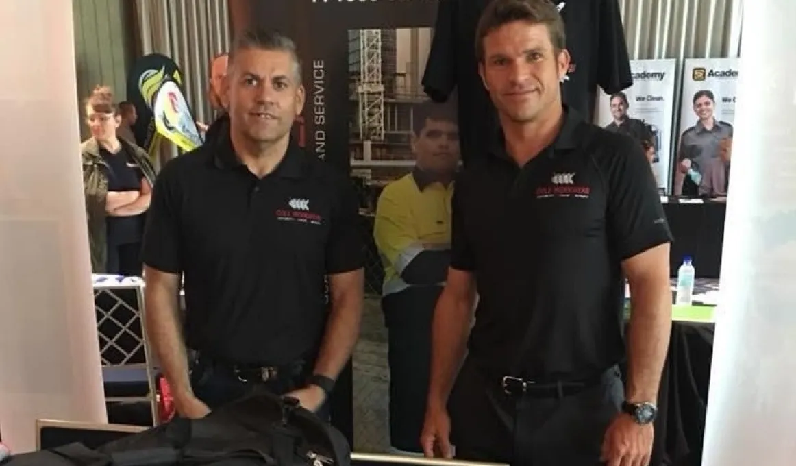 Two men in dark shirts stand facing camera. In the background are posters and a woman with arms folded.