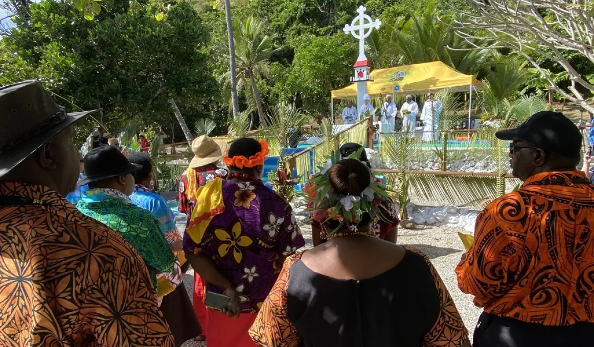 Group of people in colourful clothing stand to watch a small group on an uplifted are next to a large white cross. Behind that is a temporary gazebo with a yellow top.