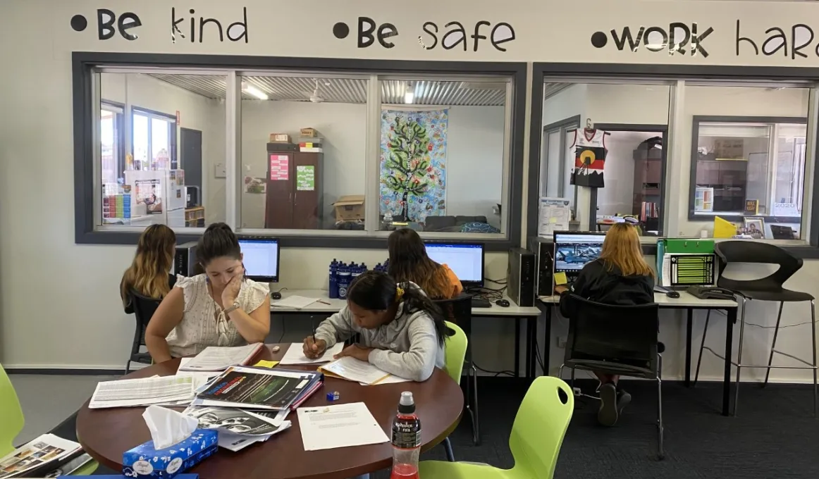 Four young women and an adult woman sit at computer desks or a table in a carpeted room with pale coloured walls and windows through to another room. Above the windows are the words: Be kind, Be safe, Work hard.