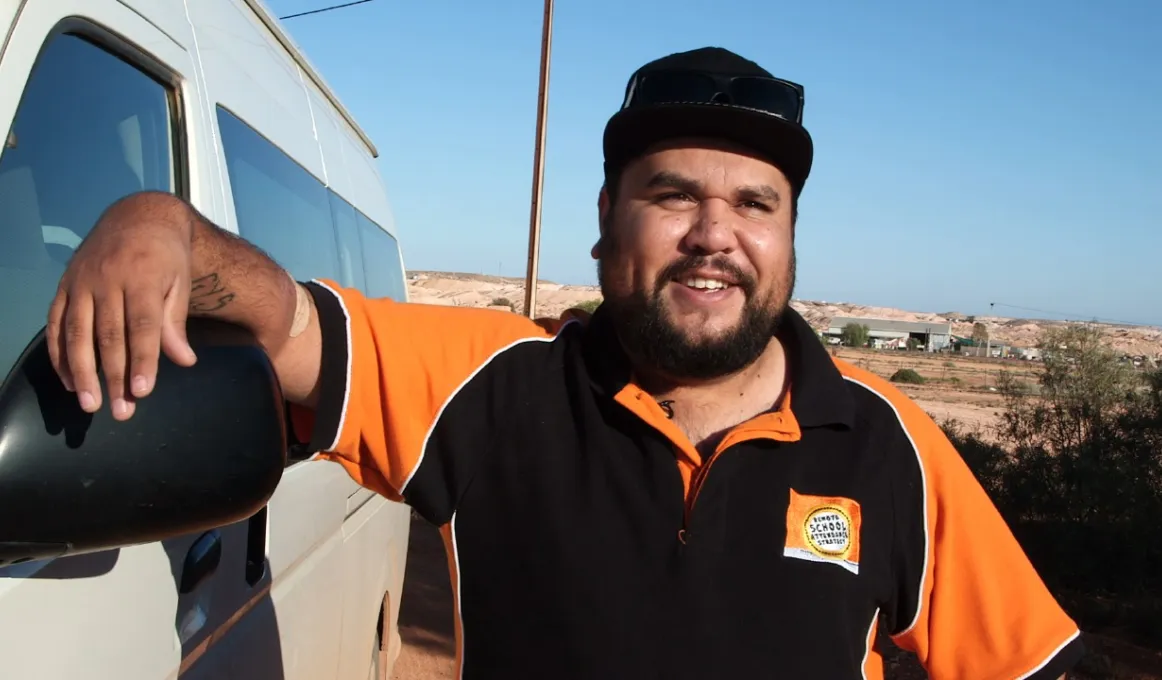 Indigenous man leaning up against a white van with buildings in the background.