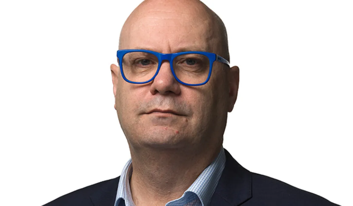 AIATSIS CEO, Craig Ritchie, posing in front of a white background with blue framed glasses, light blue shirt and navy blue blazer.