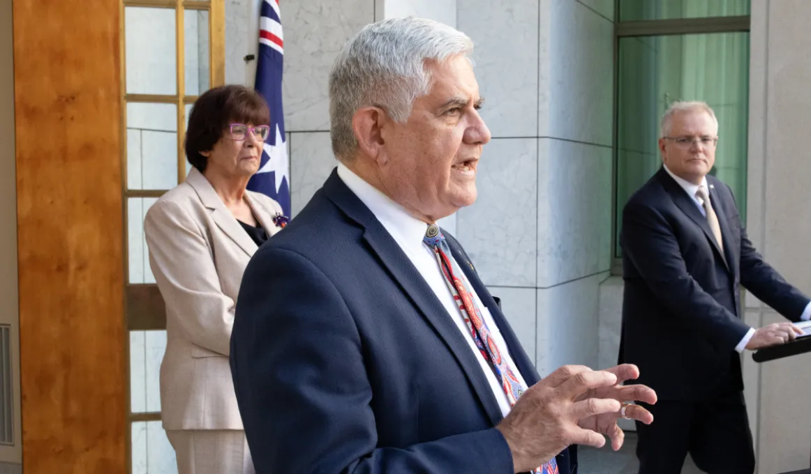 A mature aged man in blue suit with hands in front stands at centre. To the left and behind him is a mature aged woman in beige coloured wear. To the right is a mature aged man in a dark suit. He leans on a pulpit. In the background is a granite wall.