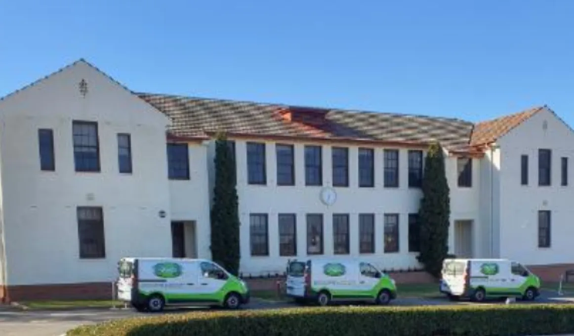 Three white and green vans line up on the roadway in front of a large white building with lots of windows and a red tile roof.