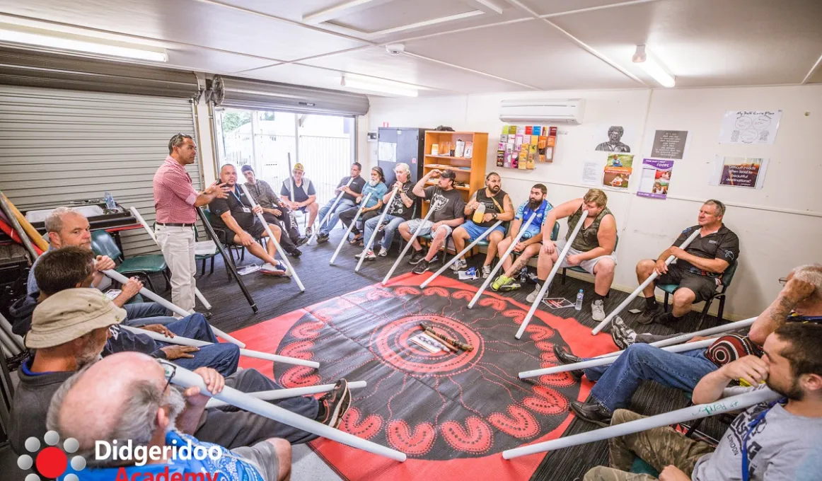 A group of men sit in a circle on chairs in a well-lit room. Each holds a white pipe pointed at the floor on which is a red and black covering. On the walls are images, a book shelf and other items. There are two roller doors on one wall.