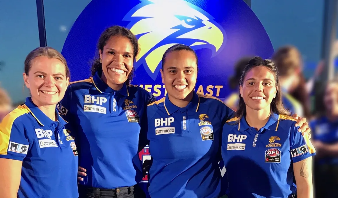 Four Indigenous women in blues shirts stand shoulder to shoulder. They wear blue and yellow shirts. Behind them is a sign with a logo featuring an eagle’s head.