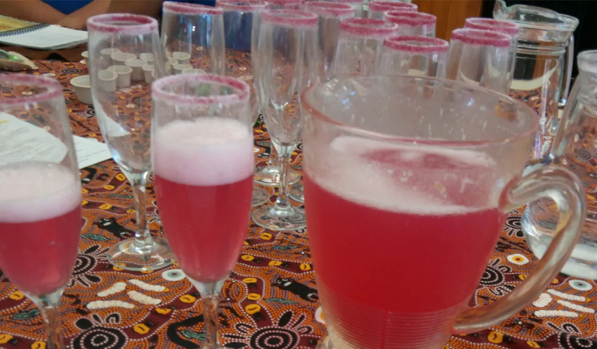 Several glasses and a jug sit on a tablecloth of Indigenous design. Some glass are empty while the jug and other glasses are filled with a red drink with a white head.