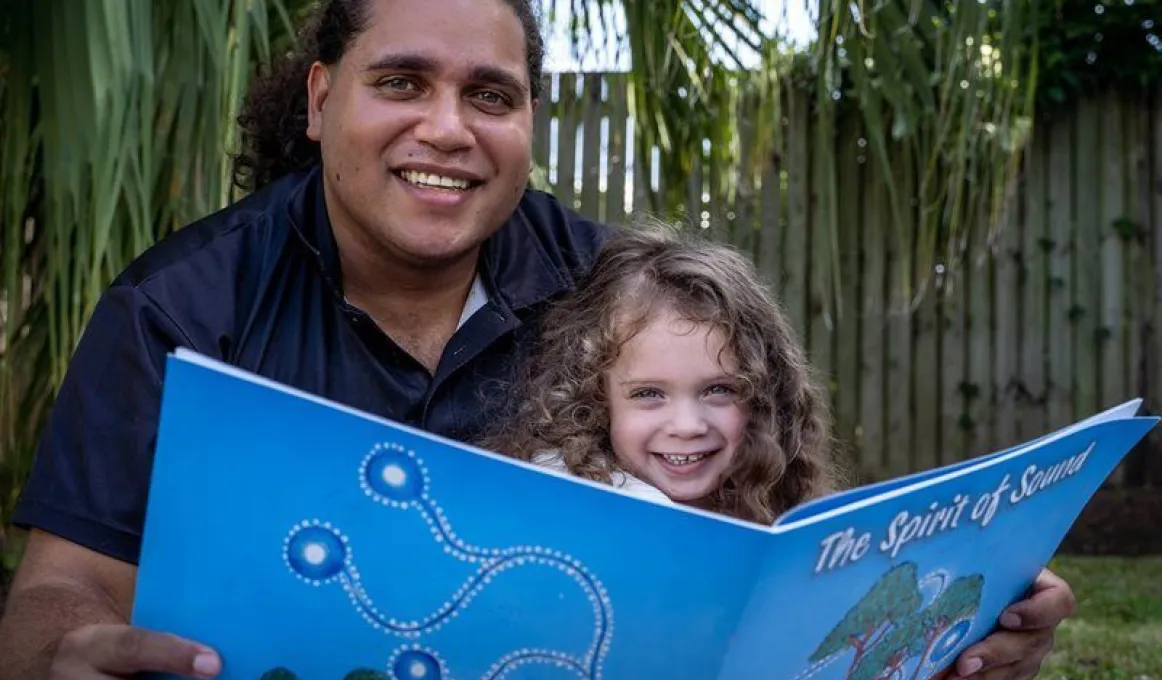 A man and a young girl hold a large blue book between them. In the background is a fence and tree branches.