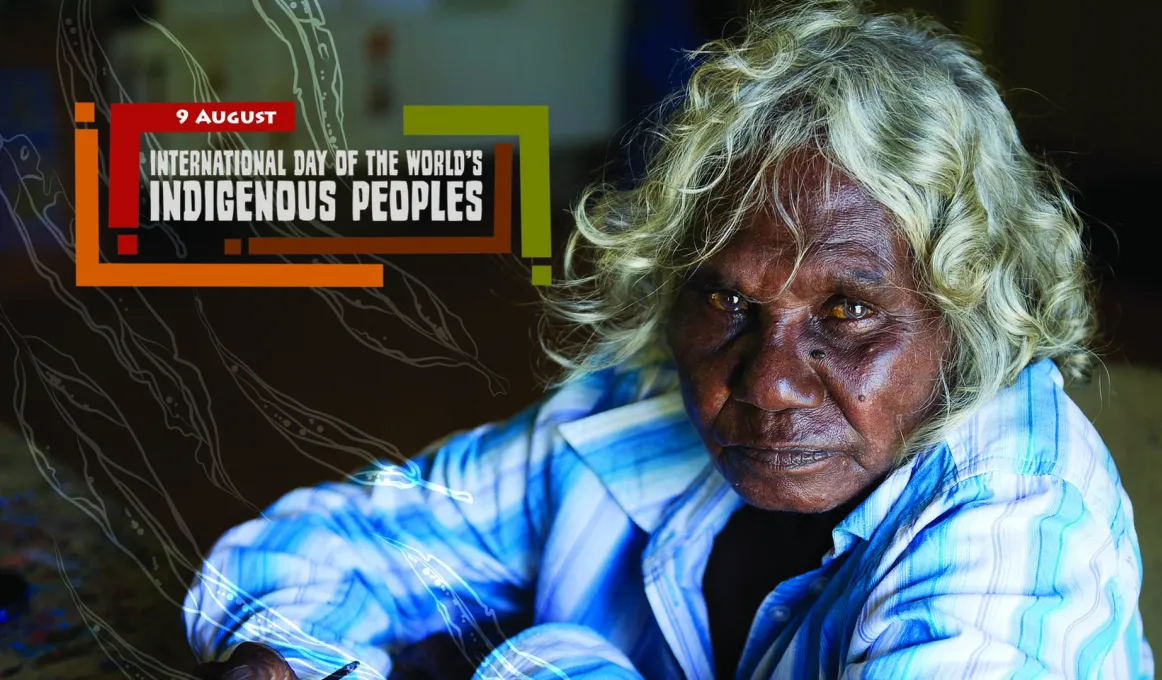 White haired Aboriginal woman wearing blue and white collared shirt sits on a floor. To her left is a graphic of leaves and the following words: 9 August International Day of the World’s Indigenous Peoples.