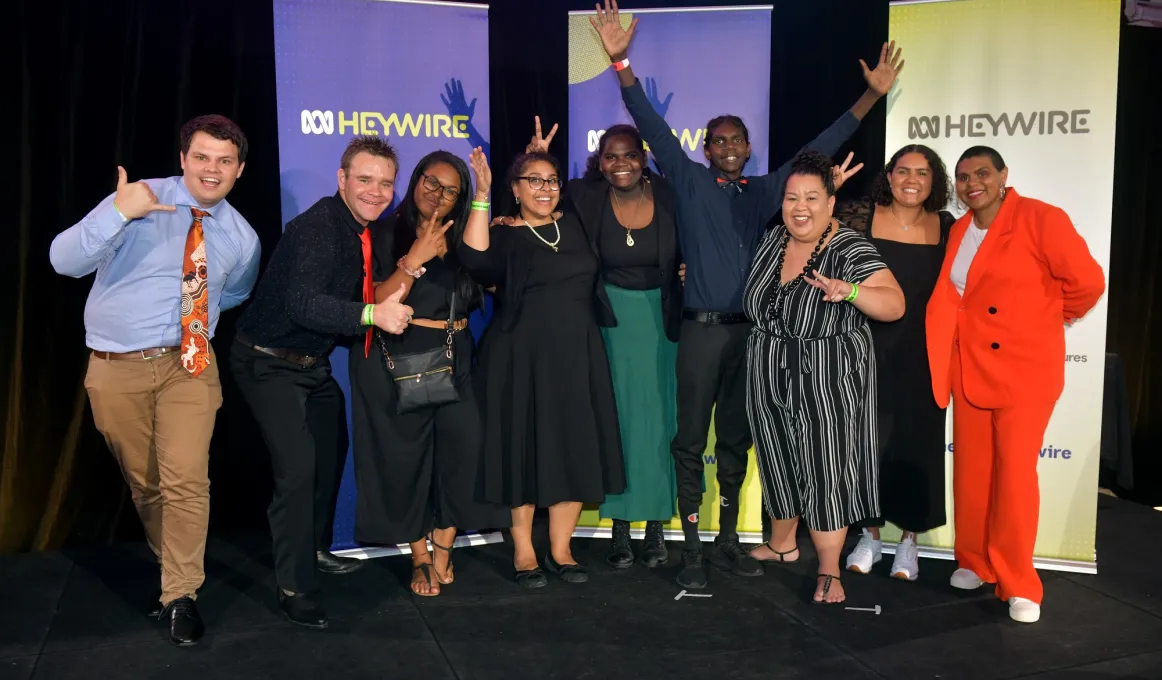 Group of 9 casually dressed Indigenous young adults stand on a black floor in front of three banners which display a logo and the word Heywire.